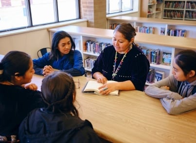 A reading group in a library setting where an adult is leading a small group of children gathered around a wooden table. The adult in the center is reading from an open book while several children of different backgrounds attentively listen. 