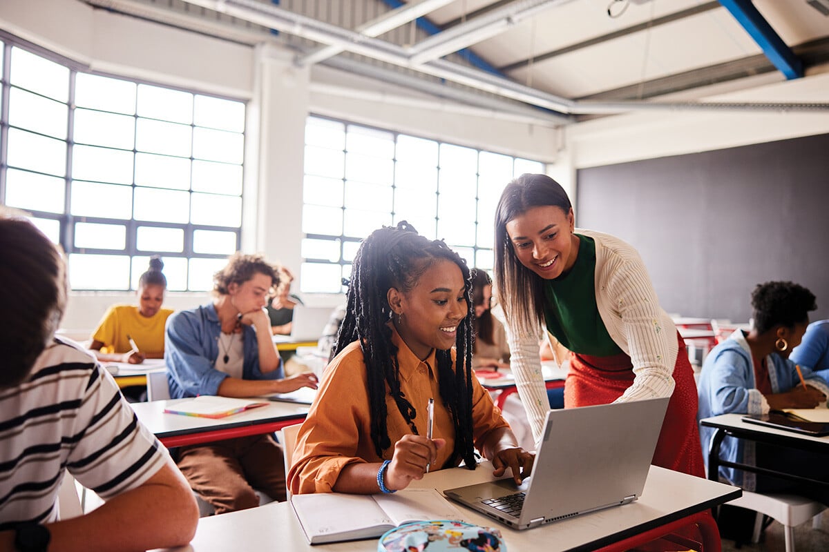 A diverse group of students collaborating in a modern classroom. In the foreground, a young woman with braided hair wearing an orange sweater is focused on a laptop while another woman in a cream cardigan and red pants leans over to help her. Other students are visible in the background working independently and in groups.