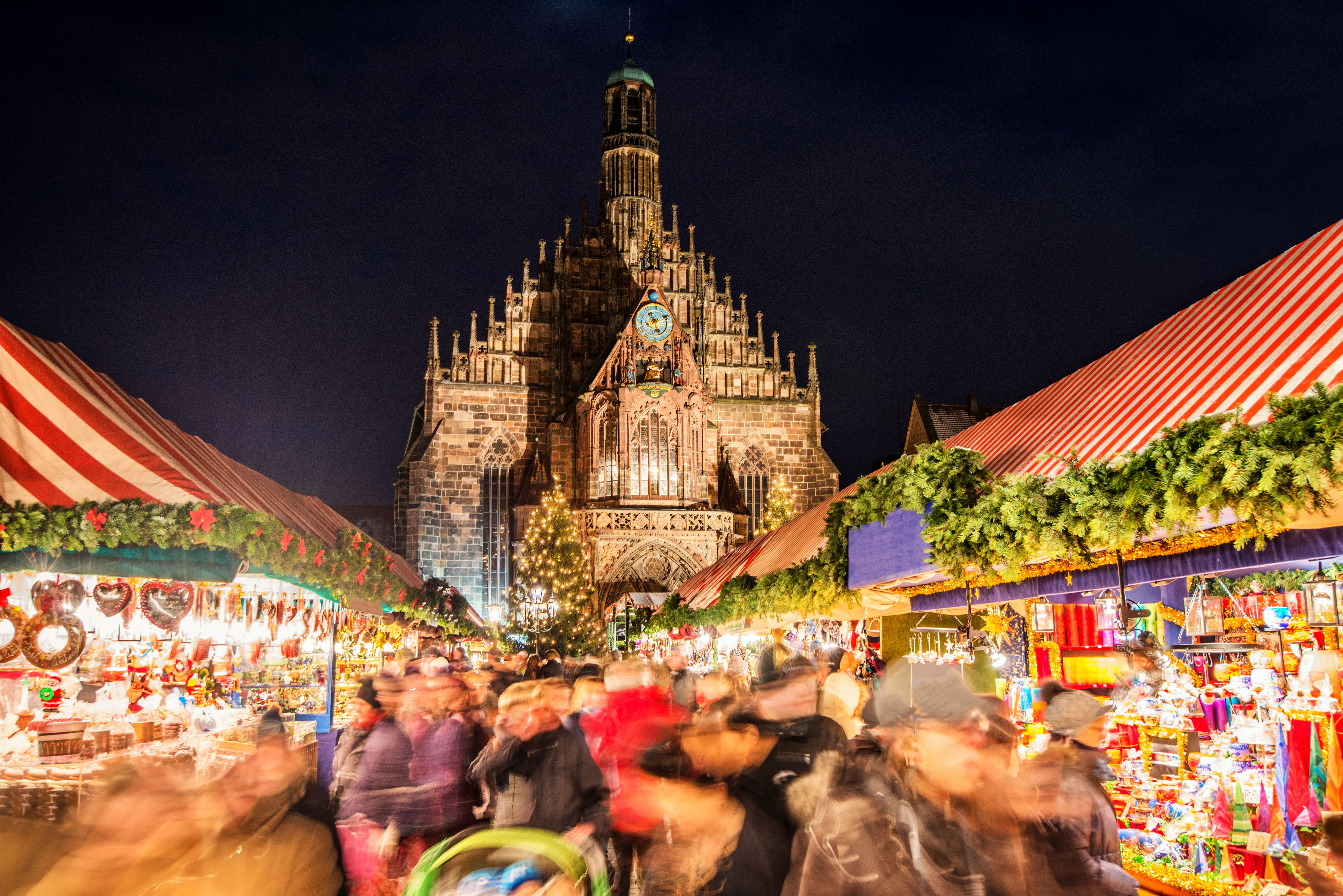 A nighttime scene of the Nuremberg Christmas Market (Christkindlesmarkt) in Germany. The beautiful Gothic Frauenkirche (Church of Our Lady) with its distinctive spires is illuminated in the background, while the foreground shows a bustling market with red and white striped stalls decorated with evergreen garlands. 