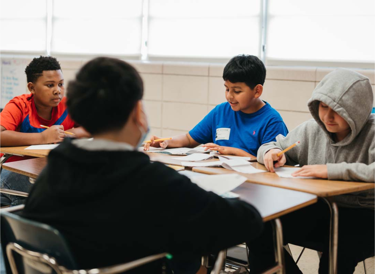 Four students sitting at classroom desks working on papers or assignments. Three students are visible from the front - one wearing a red shirt, one in blue, and one in a gray hoodie. Another student's back is visible in the foreground. 