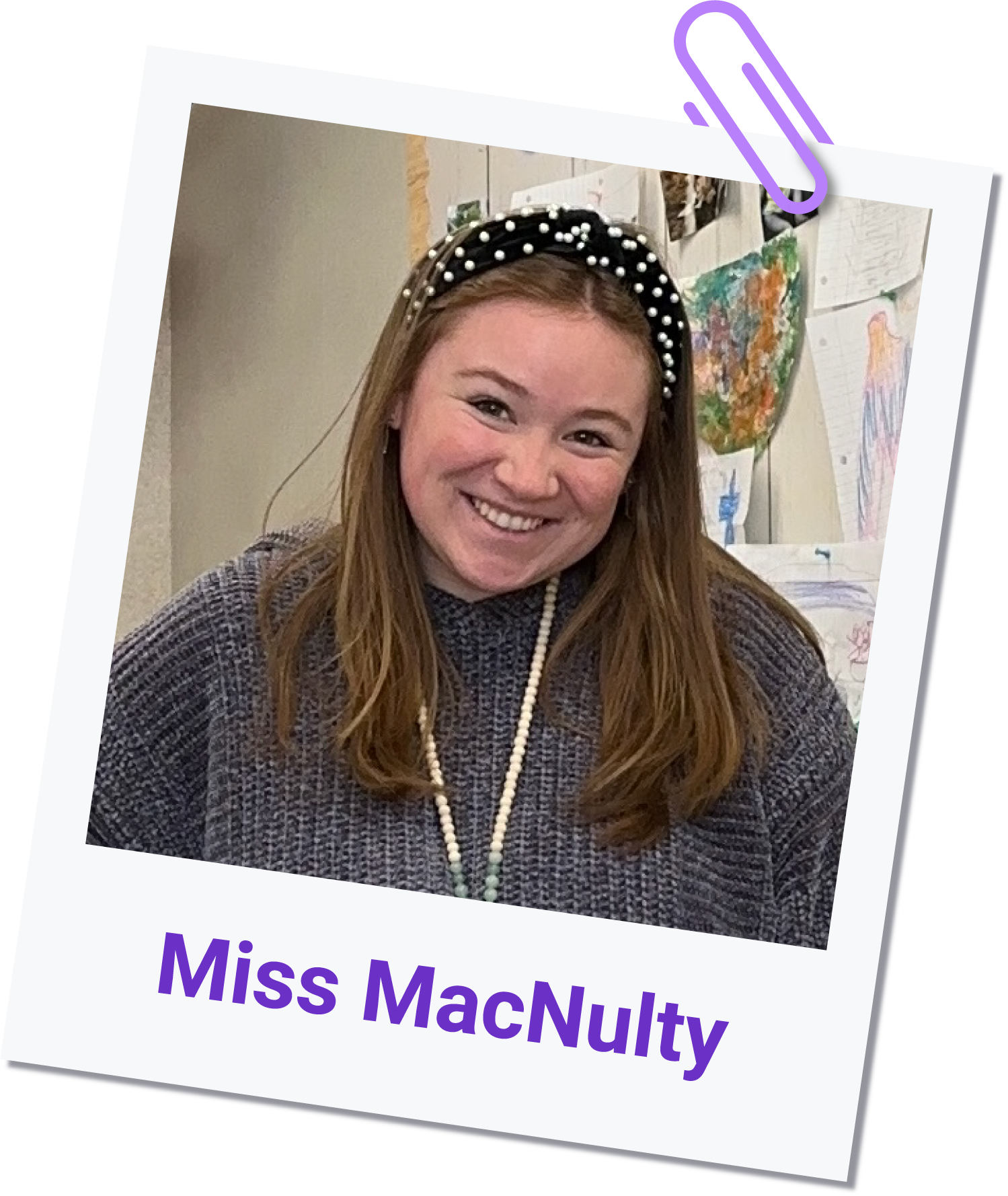 Miss Jenna MacNulty smiles at the camera, wearing a dark knit sweater and a beaded headband. She stands in front of a wall adorned with childrens drawings. Miss MacNulty is written below the photo in purple text.
